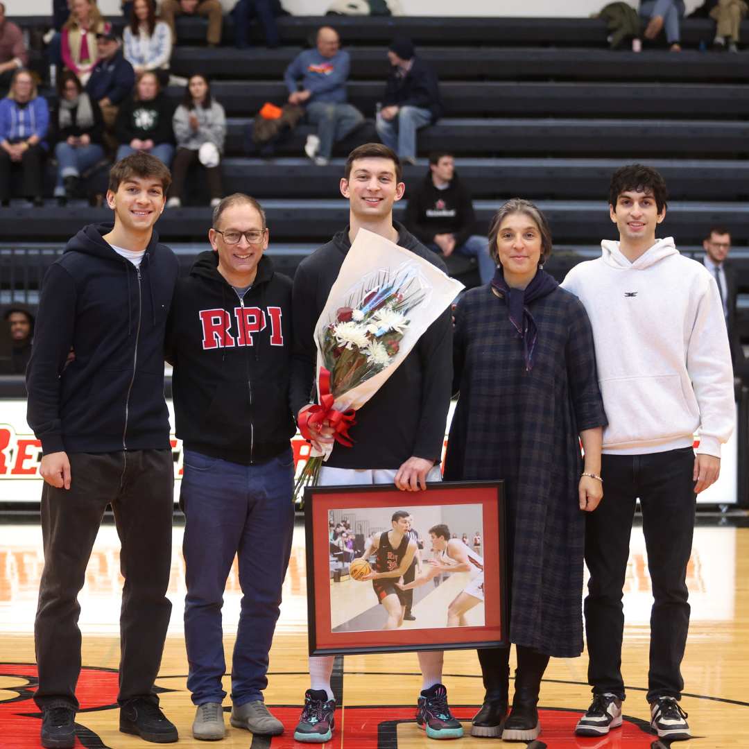 A group of five people standing together on a basketball court. The person in the middle is holding a bouquet of flowers and a framed photograph, while smiling. One person is wearing a hoodie with "RPI" printed on it. Spectators are visible in the background.