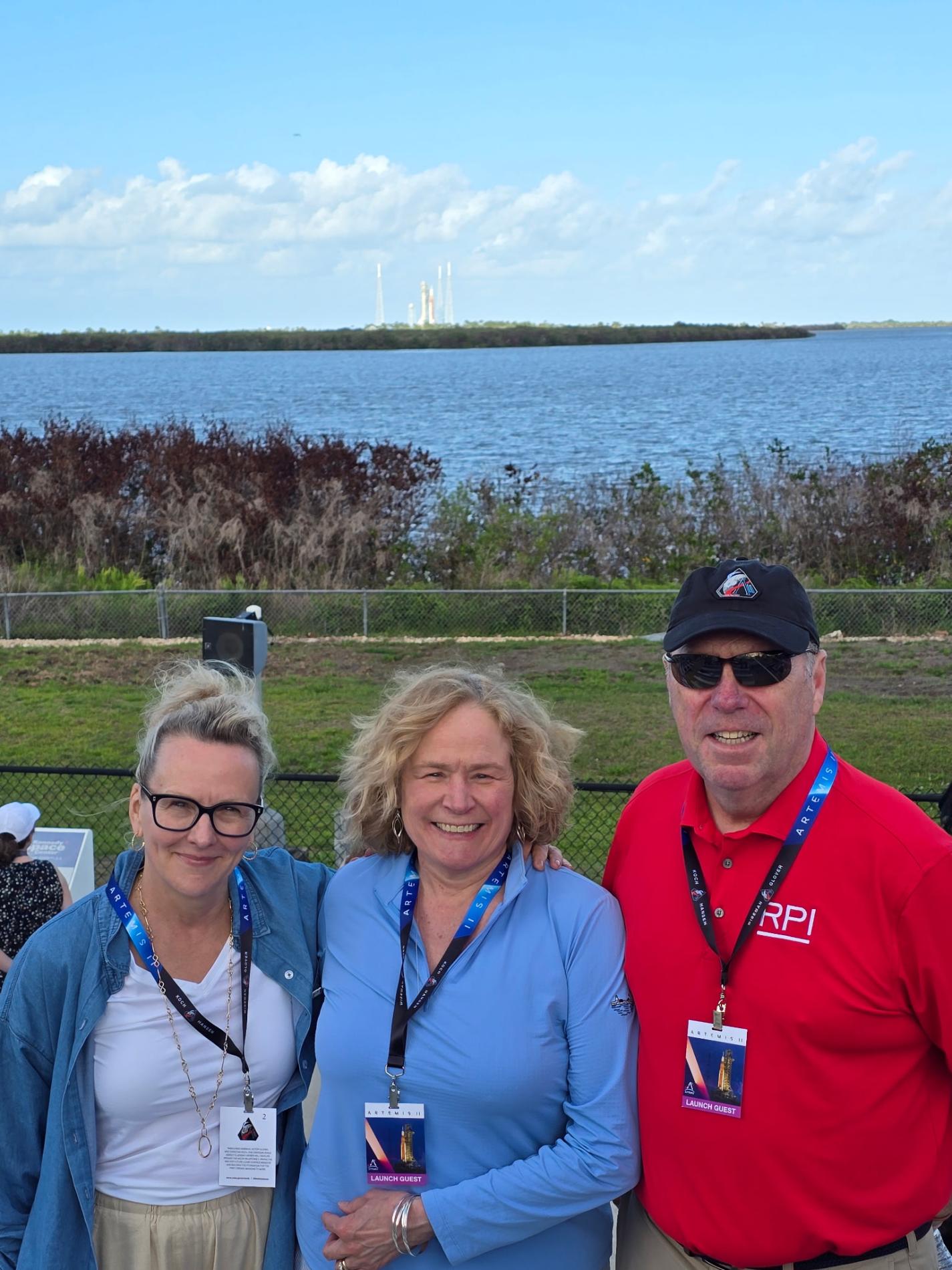 Provost Doerge, Lyn Schmidt, and President Martin Schmidt are standing outdoors near a body of water. In the background, a launch pad with a visible rocket is seen across the water. 