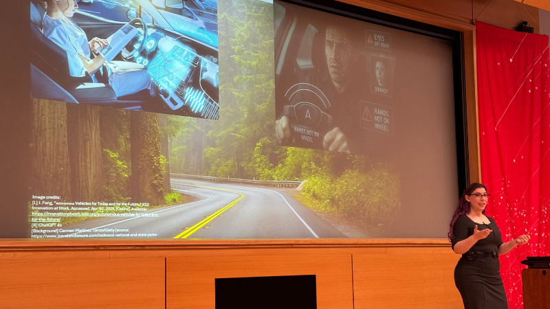 A presenter stands on stage in an auditorium, gesturing while speaking beside a large projected slide. The slide shows images of a driver interacting with advanced in-car technology interfaces.