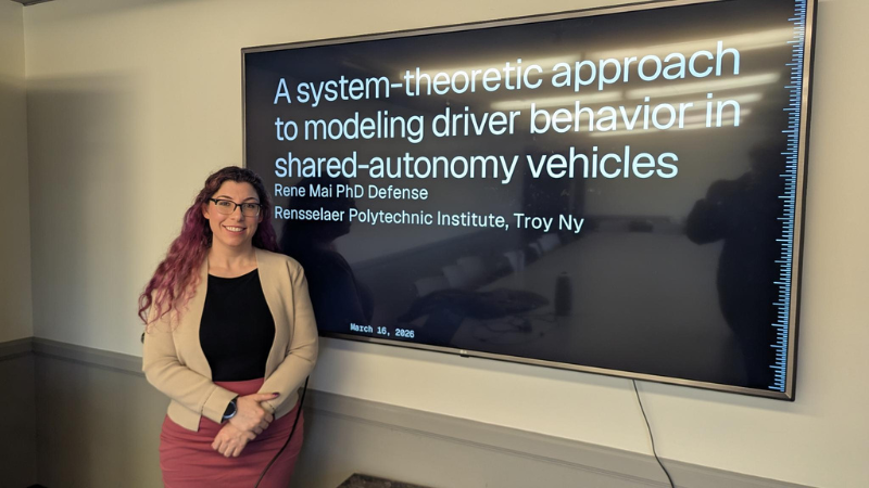 A person stands beside a large presentation screen displaying the title "A system-theoretic approach to modeling driver behavior in shared-autonomy vehicles" by Rene Mai, PhD Defense, at Rensselaer Polytechnic Institute, Troy, NY. The date shown is March 16, 2026.