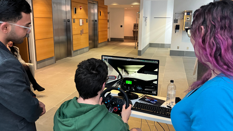 Three people gather around a driving simulator set up on a table in a building lobby. One person sits holding a steering wheel controller while looking at a monitor displaying a virtual driving scene, as two others stand nearby observing.