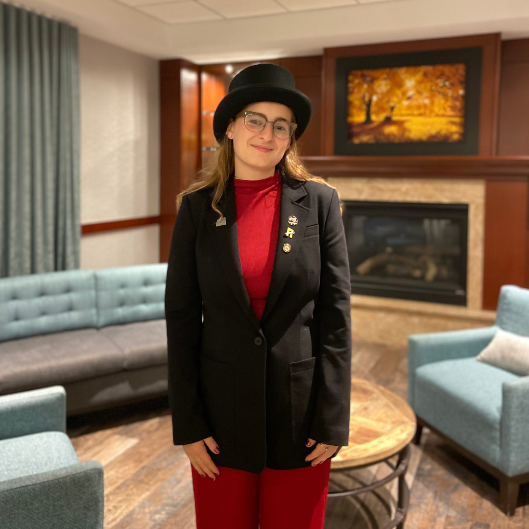 An individual wearing a black hat, glasses, a black blazer with pins, and a red turtleneck and trousers, smiling.