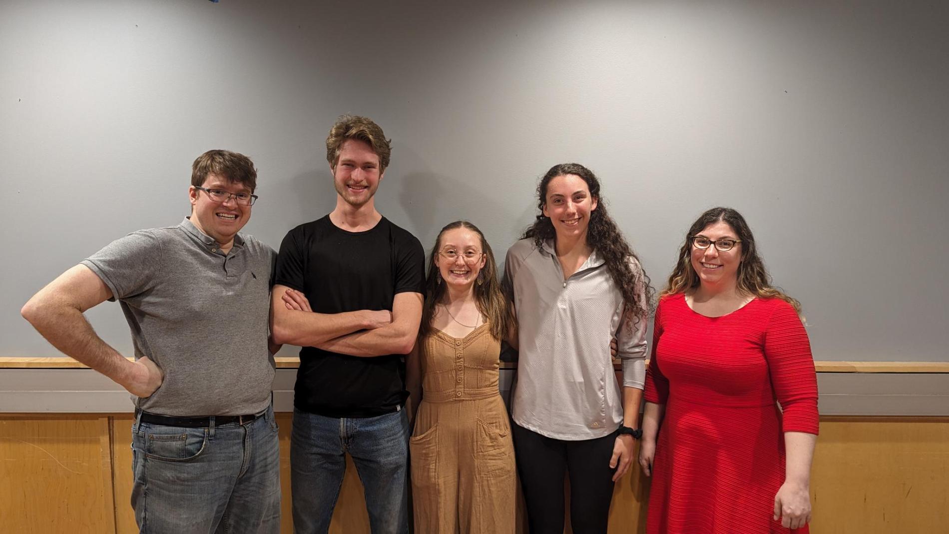 A group of five people standing side by side, smiling in a room with a plain gray wall. They are casually dressed, and the person in the center is wearing a tan outfit.