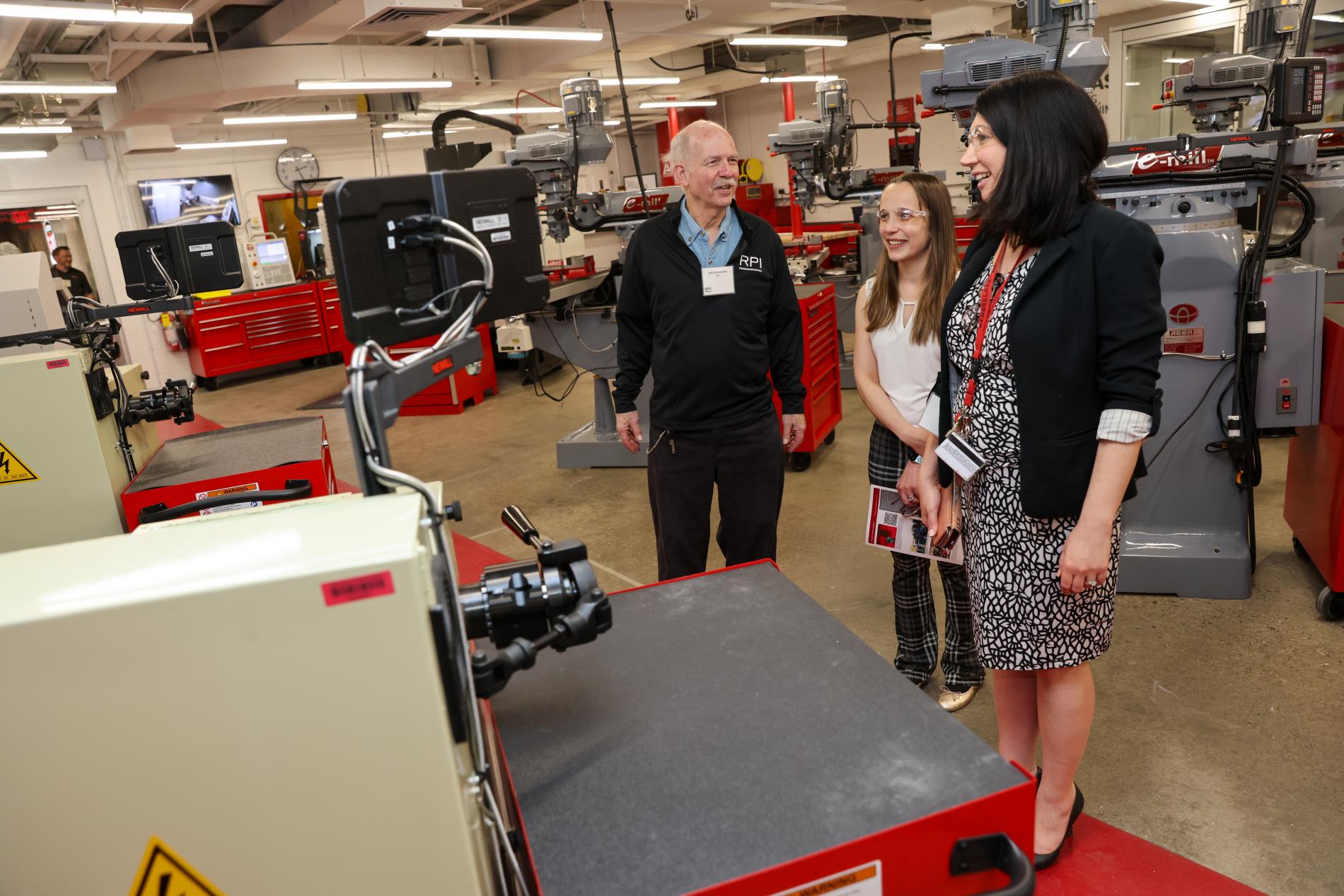 Three people are standing in a workshop area with machinery around them. They appear to be engaged in conversation. The room has red tool chests and various equipment.