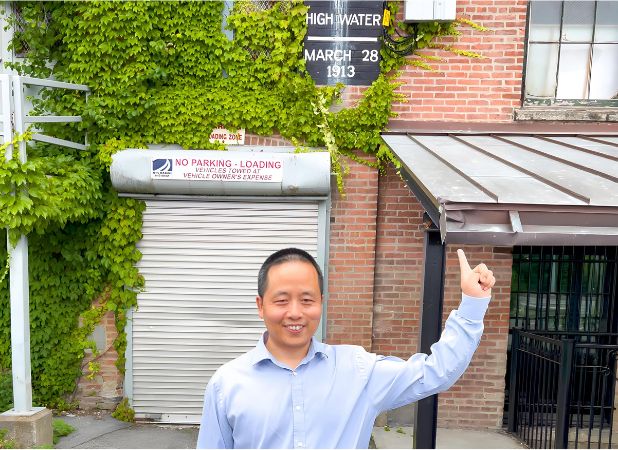 Jianxi Gao smiling and pointing upwards at a historical high water mark dated March 28, 1913, on a building covered with green ivy.