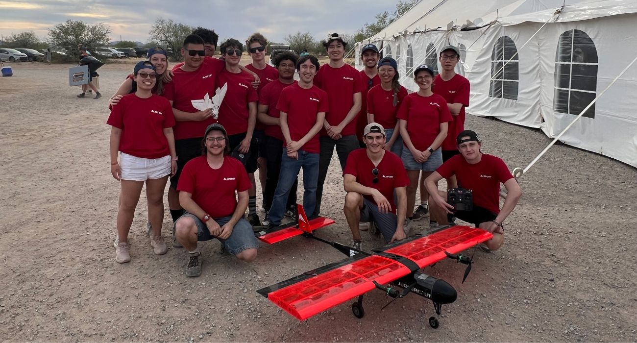 16 DBF team students wearing matching red t-shirts standing together outside with their aircraft in front of them