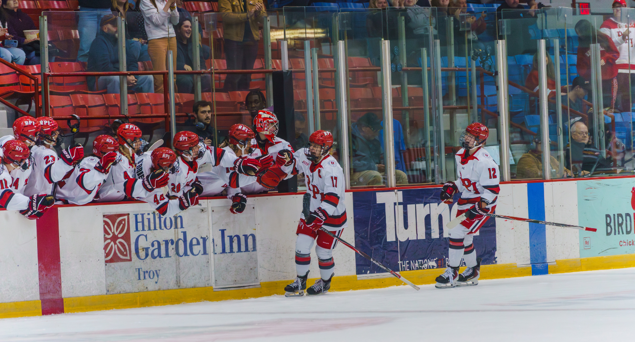 A group of RPI hockey players in red and white uniforms celebrating on the ice near the boards, with spectators in the background at an arena.