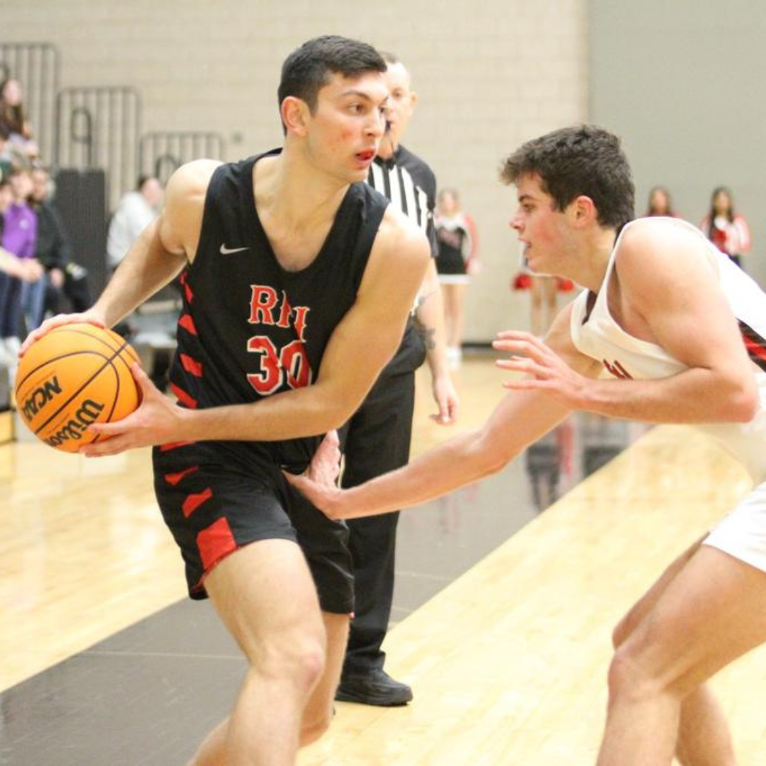 Two basketball players on a court. One player in a black and red uniform is holding a Wilson basketball, while the other player in a white uniform is defending. The referee and spectators are visible in the background.