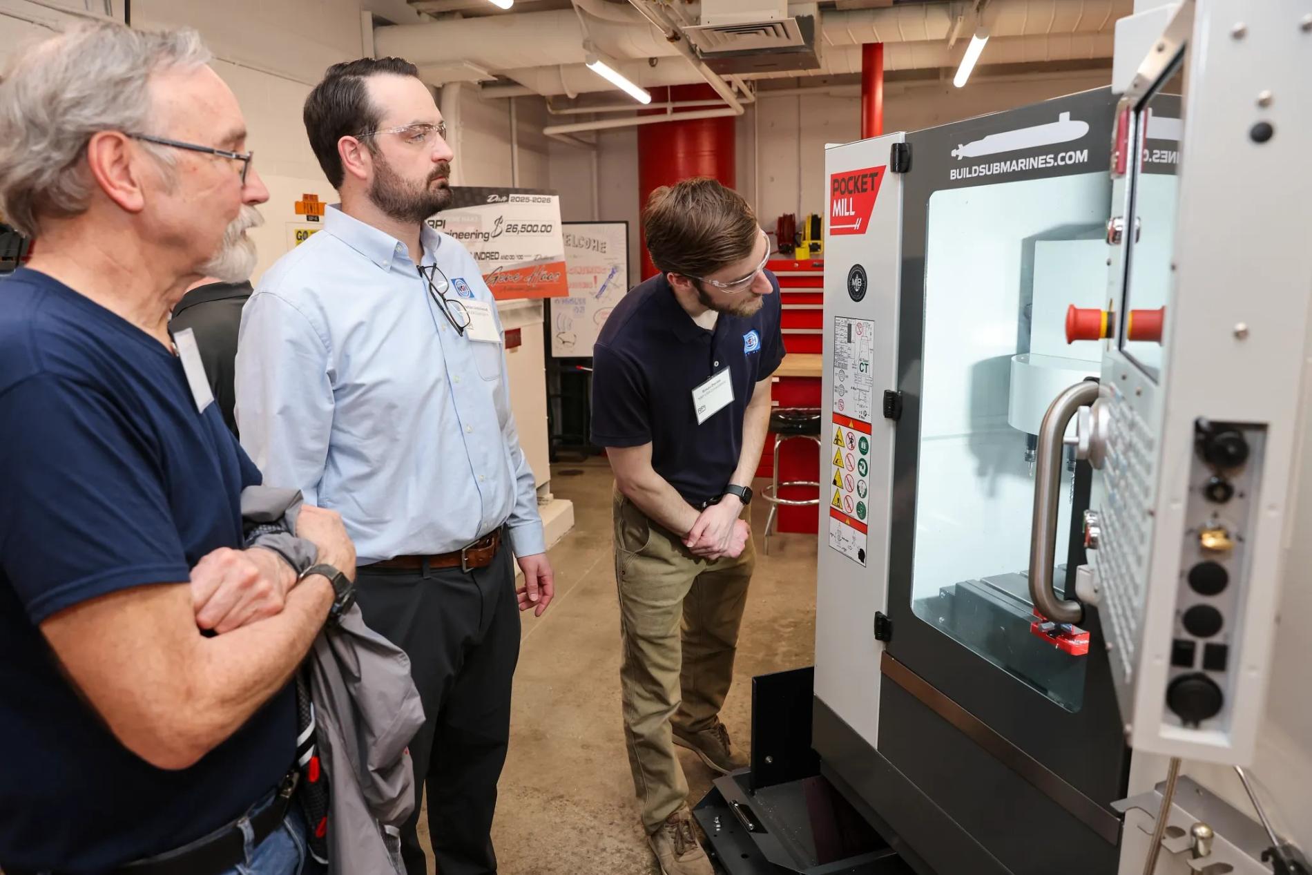 A group of three individuals stand in front of a Pocket NC machine in a workshop setting. One person is actively pointing and demonstrating the machine’s functions while the others observe attentively. Posters and industrial equipment are visible in the background.