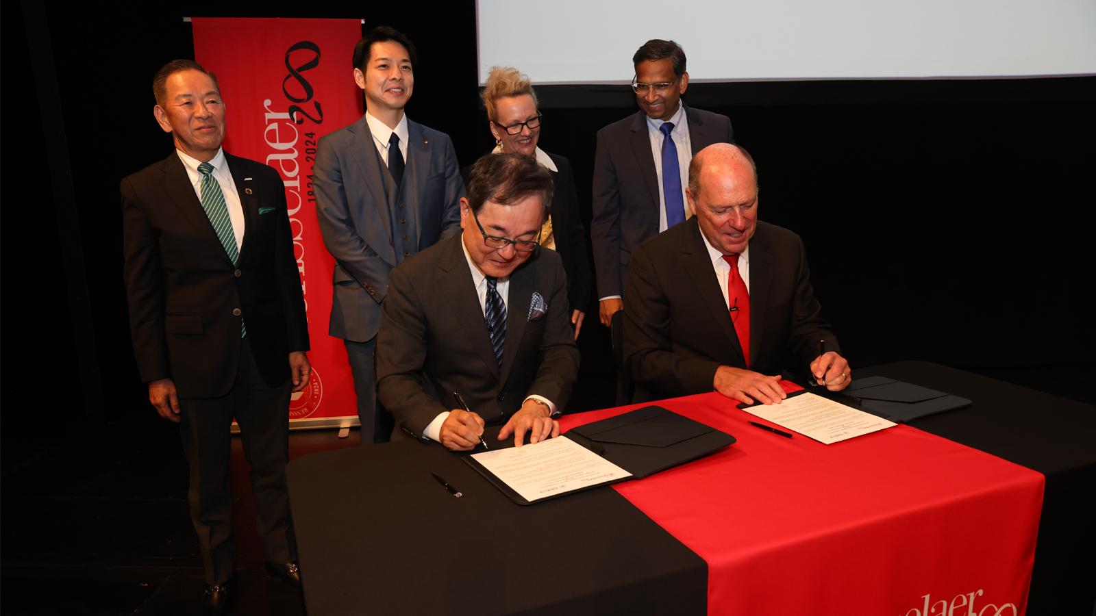 Two people sign a document with four people standing behind them