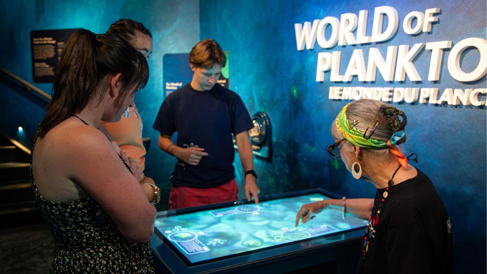 Three people interact with a digital interactive display table at the "World of Plankton" exhibit.