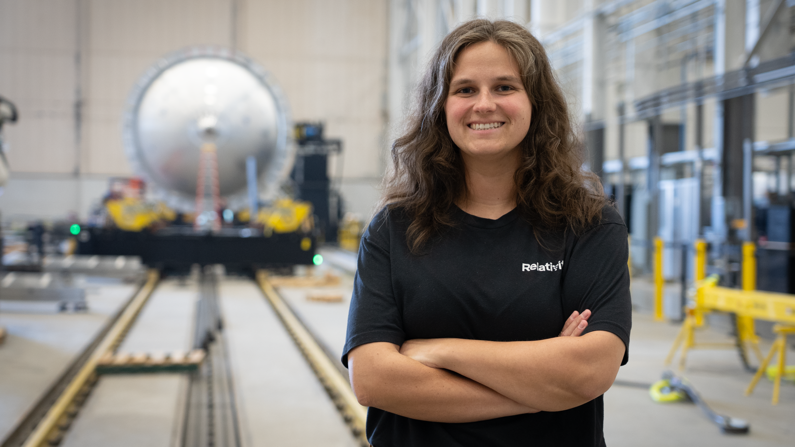 Severance standing with arms folded at her chest, smiling, standing in front of manufacturing equipment at Relativity
