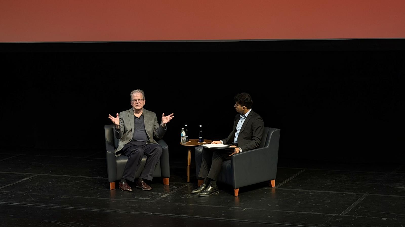 Curtis Priem and Aashrut Jain sit beside each other in chairs on stage, mid-conversation.