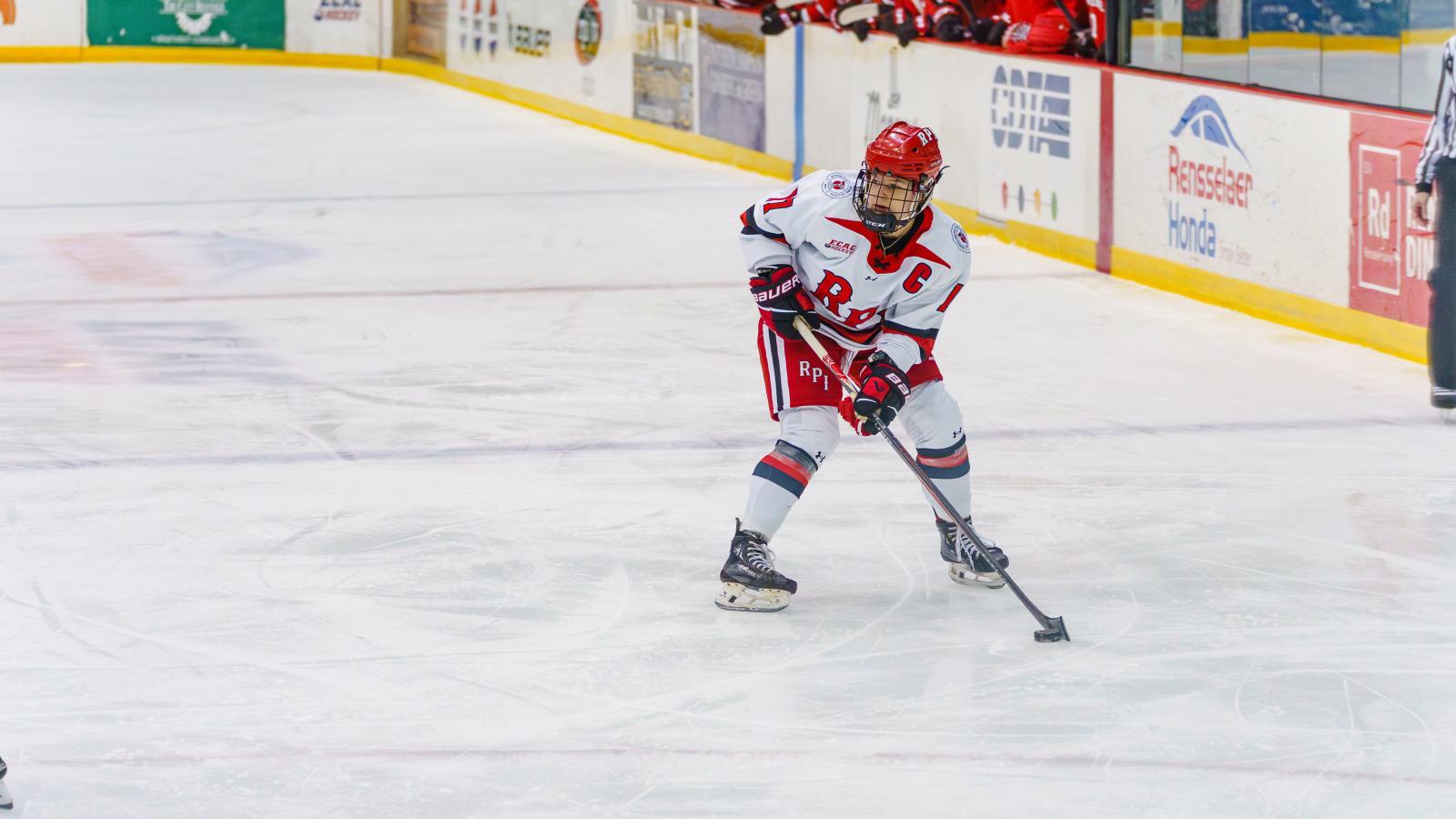 Nina Christof  in a RPI Engineers uniform skating with the puck during a game on an ice rink. Other players and the team bench are visible in the background.