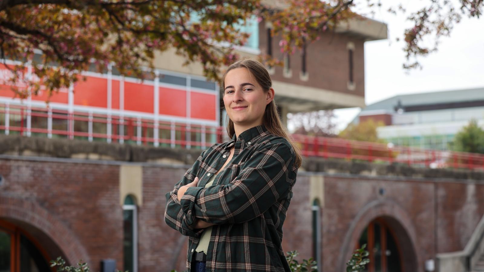 An individual with long brown hair stands with arms crossed, smiling confidently, in front of a brick archway building with red architectural accents and trees.