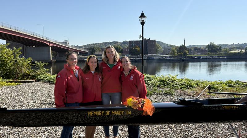 Four women standing close together, all wearing matching red jackets, behind a crew shell that reads "Memory of Mandy '04," outside on a sunny day