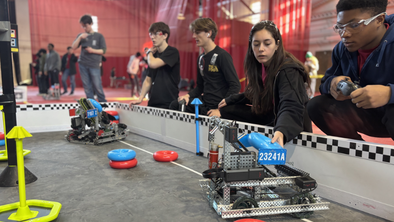 Four students participate in a robotics competition, maneuvering robots around a field with red and blue rings.