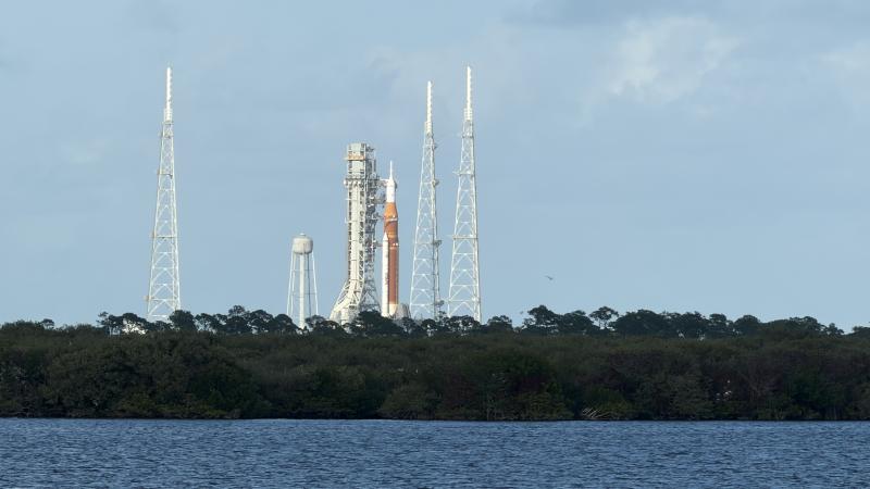 A distant view of the NASA Artemis rocket on the launch pad, surrounded by four lightning towers. The scene is set across a body of water, with a backdrop of trees and a cloudy sky.