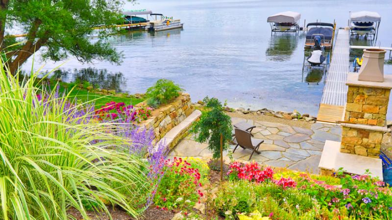 A serene lakeside scene with a stone patio and colorful flowers in the foreground. A wooden dock extends into the calm water, where several boats are moored. The background features lush greenery and a distant shoreline.