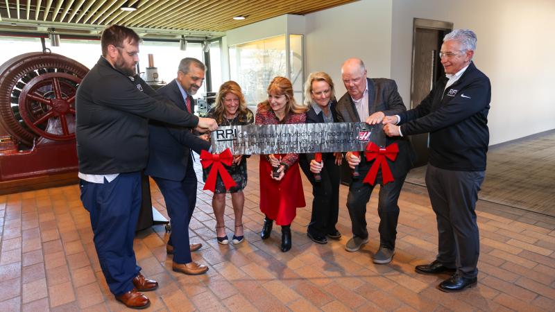 A group of people are participating in a ribbon-cutting ceremony indoors. They hold a large ribbon with the initials "RPM" and red bows attached. Behind them, there is industrial machinery visible.