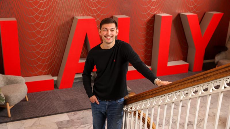 A person stands smiling in a modern indoor space with large red letters spelling "LALLY" behind. They are leaning on a decorative staircase railing.