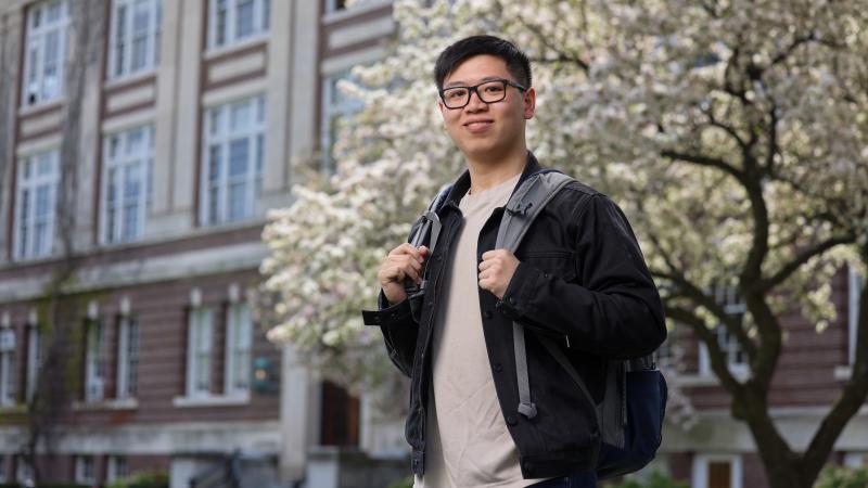 A person stands confidently outdoors, holding the straps of a backpack. They are in front of a large building and a blooming tree.