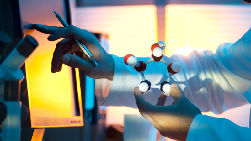 A person in a laboratory setting interacts with a computer, holding a molecular model. There are microscopes visible in the background, with warm lighting illuminating the scene.