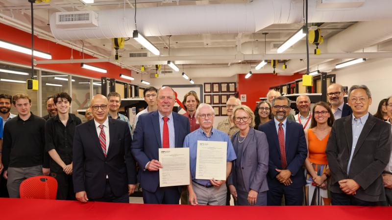 A group of people standing together in a room with red and white walls and exposed pipes. Two individuals at the front hold certificates, surrounded by others smiling.