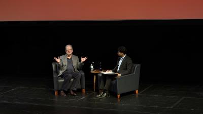 Curtis Priem and Aashrut Jain sit beside each other in chairs on stage, mid-conversation.