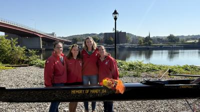 Four women standing close together, all wearing matching red jackets, behind a crew shell that reads "Memory of Mandy '04," outside on a sunny day