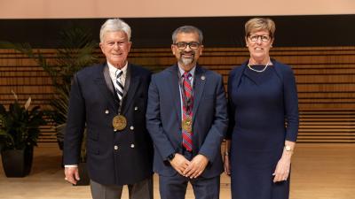 Tom Farino, Shekhar Garde, and Pat Farino standing together on stage, smiling. Tom and Shekhar are wearing medals.