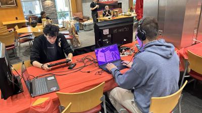 Two individuals engaged in gaming on devices at a 'Game Dev Club' event in a public setting with others in the background.