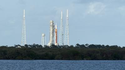 A distant view of the NASA Artemis rocket on the launch pad, surrounded by four lightning towers. The scene is set across a body of water, with a backdrop of trees and a cloudy sky.