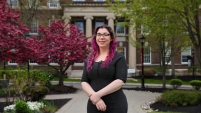 Person with brown and pink hair and glasses standing on a campus pathway in front of a columned brick building, with blooming trees and greenery.