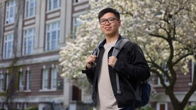 A person stands confidently outdoors, holding the straps of a backpack. They are in front of a large building and a blooming tree.