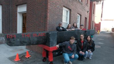 A group of people pose outdoors beside a large, handmade concrete canoe labeled "RPI The Blade." The canoe is mounted on a wheeled frame. They are near a brick building, with some orange traffic cones visible in the background.